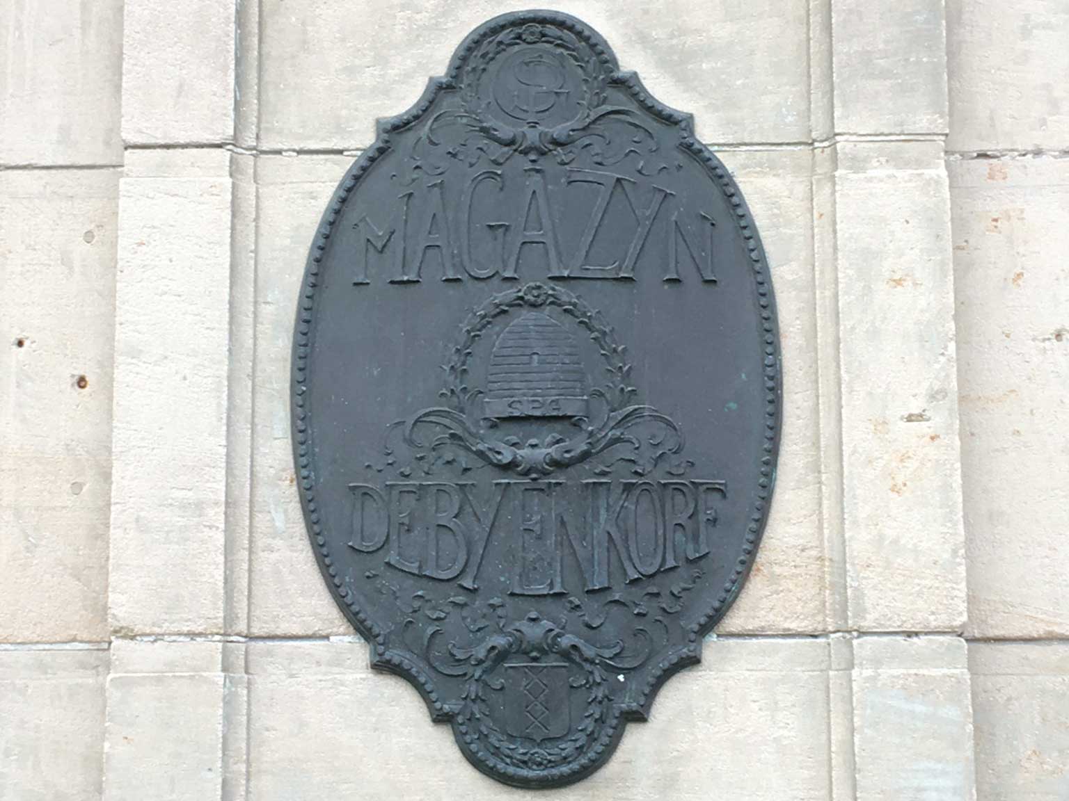 Metalen schild aan de kant van het Damrak van De Bijenkorf, Amsterdam, met bijenkorf en monogram